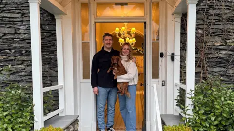 Lauren Foggo Lauren and Greg Foggo standing in front of the hotel door holding their dog, a ginger spaniel.