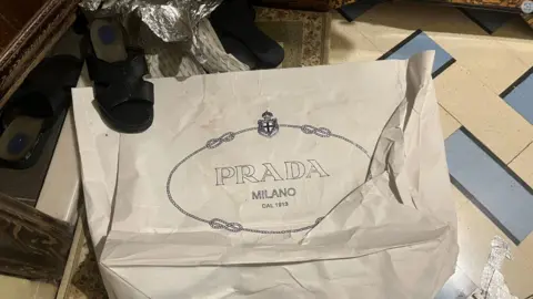 A white carrier bag with the words Prada Milano on it, with a sandal on top of one corner of the bag