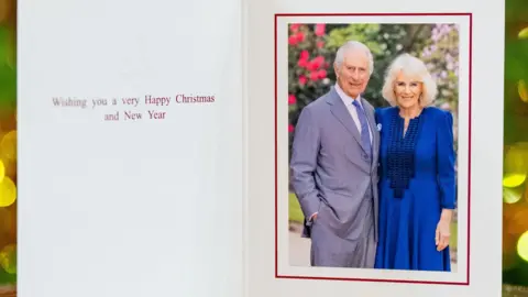 PA Media King Charles, in a grey suit, and Queen Camilla, wearing a long blue dress, are pictured in front of some shrubs in a Christmas card bearing the message: Wishing you a very Happy Christmas and New Year.