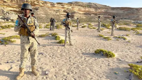 Hassan Lali / BBC Puntland Maritime Police Force officers standing in uniforms and with guns on a beach in front of cliffs in Eyl, Somalia