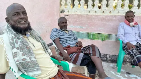 Hassan Lali / BBC Three men - two with beards - sit on plastic chairs outside in Eyl, Somalia