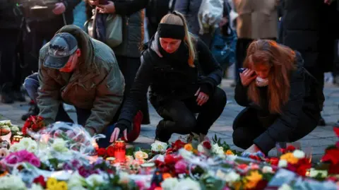 Reuters Three people - a man and two women - place tributes close to the scene of the attack. In the foreground can be seen flowers of various types and colours as well as candles in glass holders.