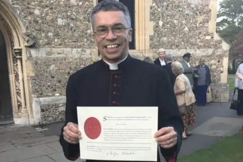 Facebook/Canvey CofE David Tudor holding up a certificate and smiling at the camera. He has thin-rimmed glasses on and is wearing black clerical clothing.