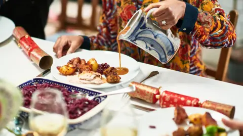 Getty Images A person wearing a orange jumper with red patterns pour gravy on to a whie plate. The plate has roast potatoes, turkey, and pigs in blankets on it. In front of them is serving dishes and there are two red and gold Christmas crackers on either side of them.