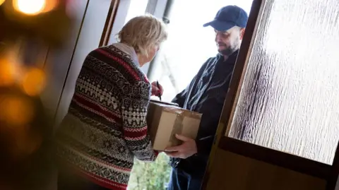 Getty Images A woman in a festive jumper that is blue, red and white is signing for a parcel at a door. A deliveryman, dressed in a black coat and baseball cap, is holding the parcel.