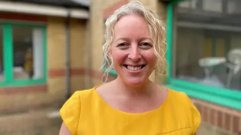 Julie has blonde, curly hair tied back and is wearing a yellow top. She is stood outdoors, with a hospital building in the background.