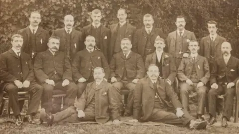 Supplied A black-and-white archive photo showing 16 men wearing dark formal suits. Seven are standing in the back row, seven are sitting in the middle row, and two are reclining on the ground at the front.