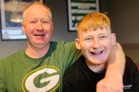 Older man smiling at camera with green T-shirt on with a large letter G on it it white with a yellow trim, with his arm round a smiling younger man - his son - smiling at camera and wearing a white T-shirt.