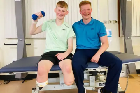 Adam Golebiewski, teenager with both legs removed below the knee, in shorts and a green T-shirt and holding a weight in his right arm, with orthopaedic rehabilitation physiotherapist Owen Cairns, in blue trousers and a blue T-shirt, both sitting on a hospital bed and smiling.