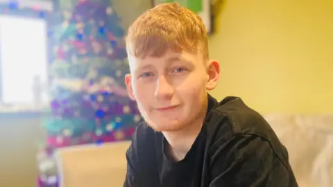 Teenage boy with ginger hair and black T-shirt smiling at camera, a decorated Christmas tree behind him in the corner.