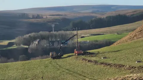 Shona Anderson A cherry picker in a field puts up a pole. A long valley stretches away into the distance.