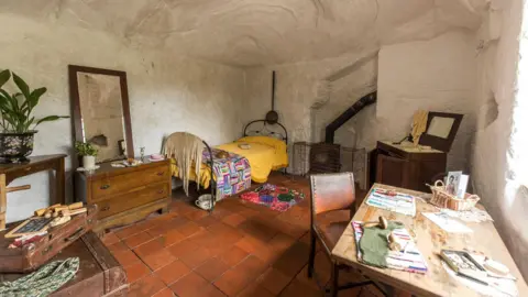 National Trust A whitewashed inside of a home with reddy tiles on the floor. An iron bed stands in one corner with a dark wood chest of drawers next to it, with a mirror on top. Other furniture including a table and chair are in the room. a pipe from a fire stove can be seen next to the bed.