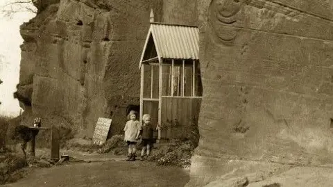 National Trust Two children by a wooden porch with a corrugated iron roof, next to a rock face. They have shorts on and are seen in sepia print. Behind them is a sign with a chalk message on it and nearby a wooden rough table with bottles on.