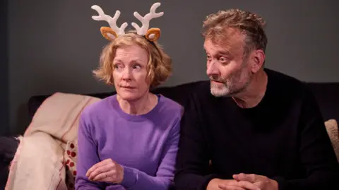 BBC/Hat Trick Productions/Adam Lawrence Right to left: Claire Skinner wearing reindeer antlers, and Hugh Dennis