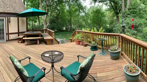 Liz Barlow A photograph of the deck shows two chairs in front of a small, metal table. A bigger table and wooden seating is located on the left under a green umbrella. Stairs lead down into the garden.