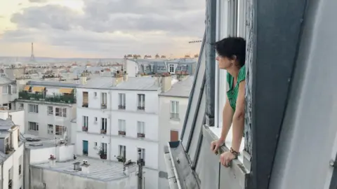 Kate Abby Kate leans out of the window as she overlooks the city's skyline. The Eiffel tower can be seen in the background on the left.