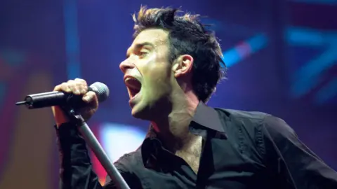 Getty Images Robbie Williams singing on stage in 2001 at Milton Keynes Bowl. He has blonde highlights in his dark hair and is holding the microphone as he sings into it.