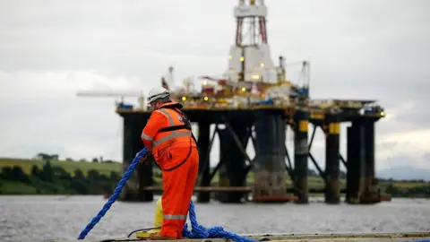 Getty Images An employee in an orange high vis suit stands on the quay side as he pulls in a rope in view of a mobile offshore drilling unit in the Port of Cromarty Firth in Cromarty