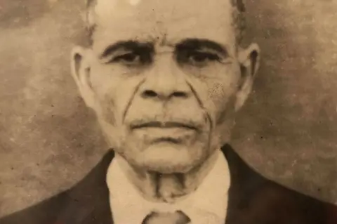 Carmen Souza's family A old sepia Carmen Souza's grandfather as a older man. He is staring straight at the camera, and is wearing a suit and tie