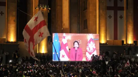 Getty Images A large crowd gathers at night in front of the Georgian Parliament building in Tbilisi, with the illuminated facade adorned with Georgian flags. A giant screen displays President Salome Zourabichvili delivering a speech, standing between the Georgian and European Union flags.