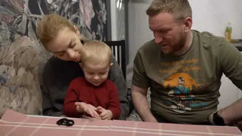 A little boy sits on a woman's lap at a table, his father next to him, and a toy car on the table