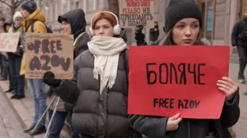 People are seen at a protest, including a young woman holding a red sign saying "Free Azov"