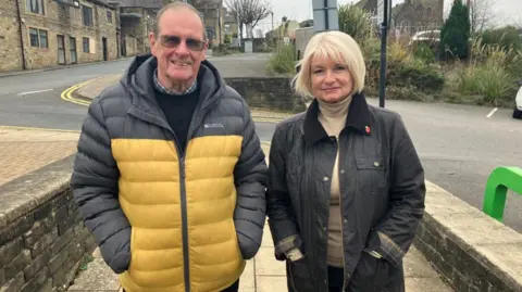 A gentleman in his seventies and a lady of similar age stand next to one another on a public street. The gentleman is wearing a yellow and black coat, while the lady is wearing a black coat with a poppy pinned to her collar. Both are smiling into the camera.