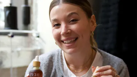 Isobel Perl Isobel, wearing a white top and grey cardigan, smiles at the camera and is holding two of her beauty products