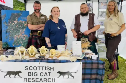 Paul Macdonald The four members of the project are at an event. They are standing next to a table draped in tartan and with a selection of big cat skulls set out on it. They also have a map showing where big cat sightings have been reported.