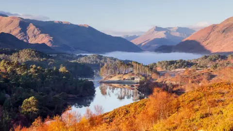 Getty Images The landscape of heather, trees and mountains are in autumn colours of yellow and gold. There is a loch with trees reflected in it.