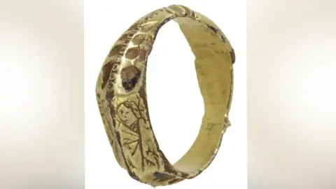 Andrew Williams/Norfolk County Council A late medieval gilded silver ring resting on its side so its plain interior can be seen. The exterior is highly decorated and the side facing the viewer shows a crude image of a woman holding a palm. 