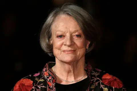 Getty Images Dame Maggie Smith photographed in 2015, aged 80, against a black background. She is smiling and wearing a black top, a simple necklace and a collar-less floral jacket.