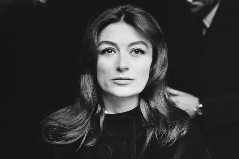 Getty Images 1960s news photo of Anouk Aimée, when she would have been in her early 30s. She is captured close-up and is staring ahead in a very confident manner. She has long dark hair and is wearing something black, which is indistinguishable from the very dark background of the photo.