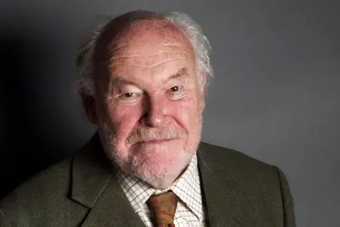 Alamy A photo of Timothy West taken when he was a fairly elderly man, standing against a grey background. He is mostly bald, with some grey hair and is unshaven. He is wearing a greyish jacket, a checked shirt and a floral tie.