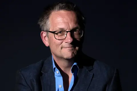 Getty Images Publicity shot of Michael Mosley against a black background. He has glasses, slightly receding greyish hair, and is smiling as he looks to the camera. He wears a dark jacket and a cobalt blue shirt with the top two buttons undone.