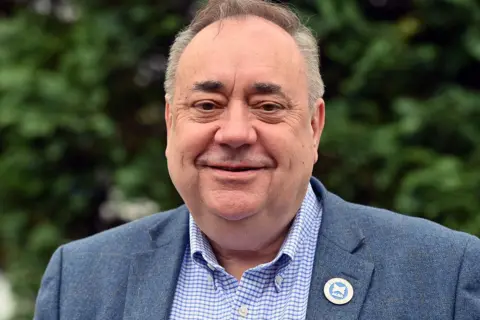 Getty Images Alex Salmond is photographed outside against a blurry background of trees. He is bald and clean-shaven, with distinctively bushy eyebrows, and smiles for the camera. He wears a light blue jacket with an "Alba" badge pinned to it, and a blue-check button-down shirt.