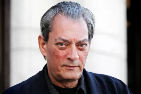 Reuters Paul Auster stares intently and seriously at the camera in this shot. He has slicked back grey hair. He appears to be outdoors and is wearing a dark jacket and dark shirt over a black T-shirt