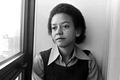 Getty Images A black-and-white photo of Nikki Giovanni in a hotel room in Chicago in the mid-70s. She is looking thoughtfully out of a window on an upper floor. She has shortish hair and is wearing a black tank top over a shirt with wing-tip collars