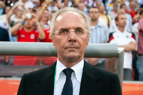 Getty Images Sven-Goran Eriksson looks impassively beyond the camera in front of a crowd of spectators in England replica shirts. He has receding grey hair and wears frameless spectacles, a black suit jacket, white shirt and dark tie.