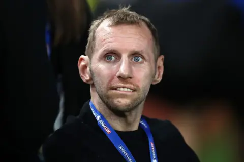 Getty Images Rob Burrow pictured during the last few years of his life when he was suffering from MND - he has receding straw-coloured hair and he is looking beyond the camera, biting his lip. He seems to be in a wheelchair being pushed by an indistinct figure, whose lower half is visible in the photo. Rob Burrows wears a black sweater, with a blue lanyard around his neck.