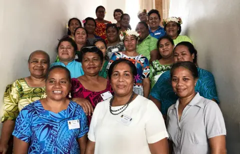 UNDP Dr Puakena Boreham, centre,  wearing white, surrounded by 20 women, all smiling, some in traditional Tuvaluan dress with flowers in their hair, in a photo from 2018