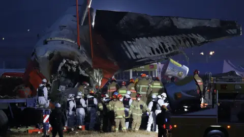 Getty Images More than a dozen firefighters and other emergency workers gather round the remains of a wrecked plane. It is night and it is dark