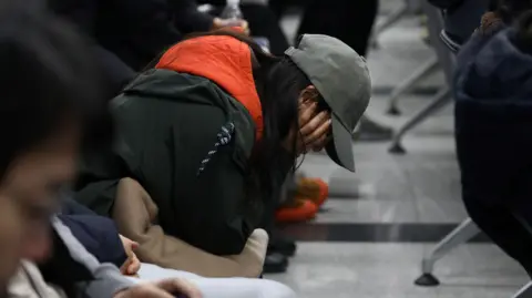 Getty Images A woman sits on a row of seats at an airport with her head in her hands. She is wearing a baseball cap and you can't see her face