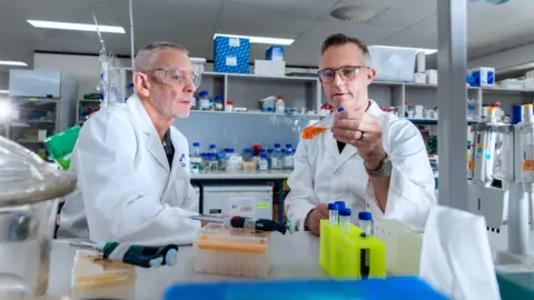 UniSC Two men examine a test tube with orang liquid inside while sitting in laboratory. They are wearing safety goggles and white coats.