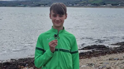 PA Media Liutauras is standing on a beach with the sea behind him smiling at the camera as he holds up his Lego black octopus