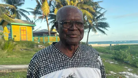 Steven Bourne, standing outdoors, has short grey hair and glasses with big black frames. He is wearing a black-and-white top with Chinese characters on it. Behind him is a yellow wooden house with palm trees nearby. Further away, beyond some scrubland, is the beach and the sea.