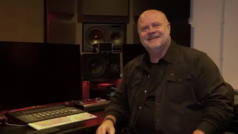 Emyr Afan, a bald man in a dark shirt, smiles at the camera as he sits at a sound mixing desk.