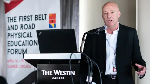 University of Worcester Mick Donovan, a middle-aged man with a shaved head, speaks into a microphone at a lectern labelled "The Westin Zagreb". A banner behind him reads "THE FIRST BELT AND ROAD PHYSICAL EDUCATION FORUM".