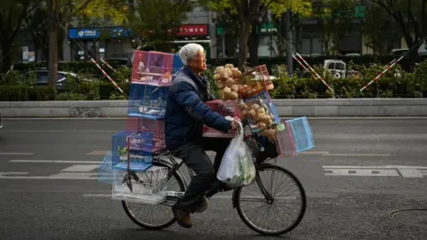 Getty Images An elderly man rides a bicycle with bird cages on a street in Beijing on November 20, 2023. Behind him are bushes. The cages are red, white and blue