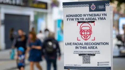 Getty Images A close up of a poster in a city centre with people walking in the background. The poster says 'Live facial recognition in this area' with a barcode and an outline of facial structure. 